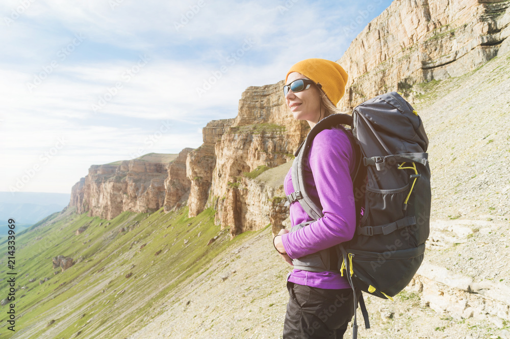 Smiling girl traveler in a yellow hat and a pair of sunglasses stands at the foot of epic rocks with a backpack next and looks away