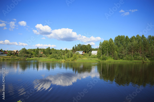 Lake reflecting sky with clouds on it flowing through a small village and covering banks with trees, shrubs and plants growing on them
