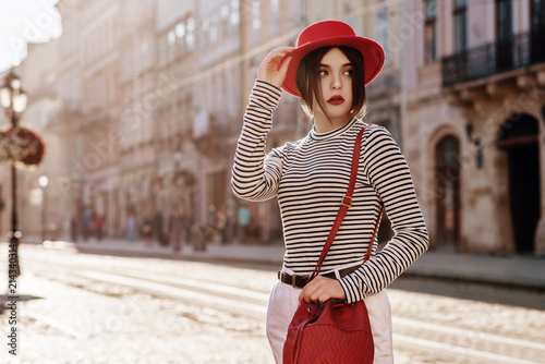 Outdoor portrait of young beautiful fashionable woman wearing stylish red hat, striped turtleneck, white jeans, holding red bag. Copy, empty space for text