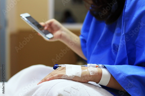 The woman patient in the hospital in blue dress in bed. Seeing the left hand was intravenous injection by dropping bottle. Using Mobile Phone.