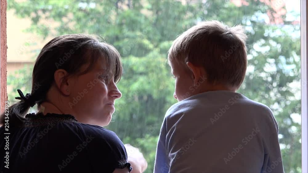 Mother and son admire summer rain at window, boy touching drops Mother and son admire summer rain at window, boy touching drops
