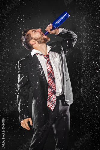 Drunk hopeless young bearded businessman drinking heavily from bottle under the rain, on black background. Social problem concept