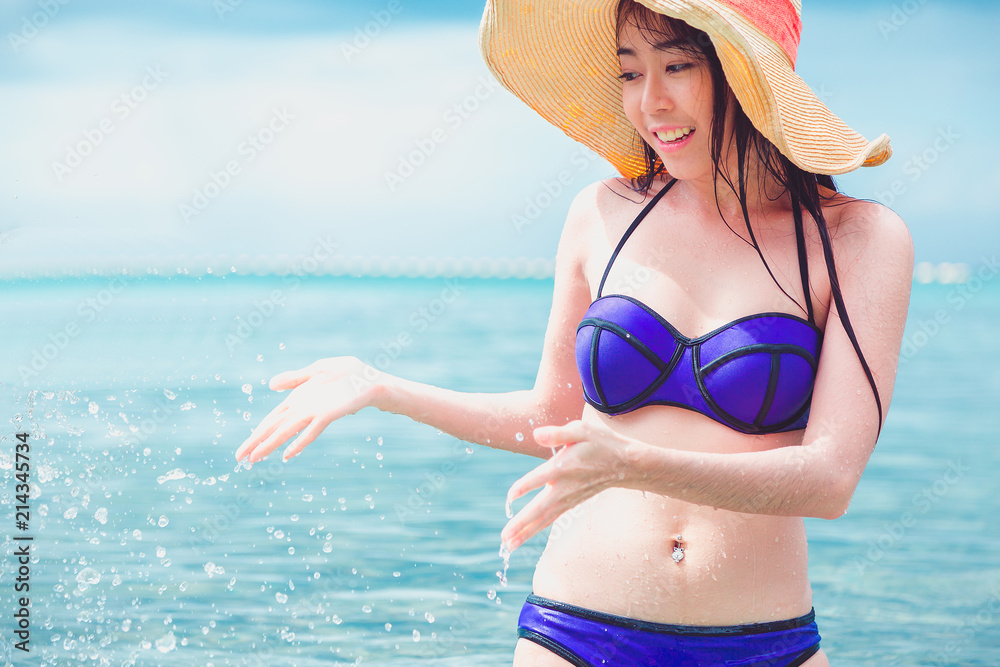 Fototapeta Beach woman happy on travel vacation holidays in bikini on by blue ocean sea at tropical resort. Cheerful smiling excited mixed race girl wearing sun hat laughing full of joy looking at camera.