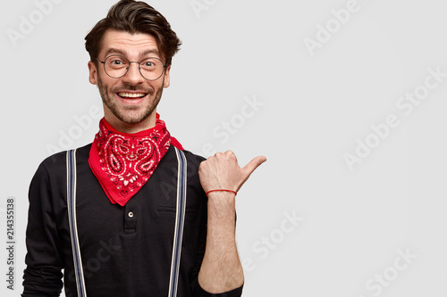 Satisfied handsome man in fashionable clothes, happy to show you direction, points aside with thumb, has joyful facial expression and charming smile, isolated on white background, free space for text