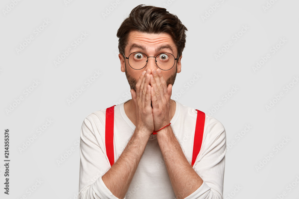 Emotive Caucasian male covers mouth with fear, exclaims in horror, sees something scarying, has trendy hairdo, wears white sweater with red suspenders, isolated over studio wall. Reaction concept Emotive Caucasian male covers mouth with fear, exclaims in horror, sees something scarying, has trendy hairdo, wears white sweater with red suspenders, isolated over studio wall. Reaction concept