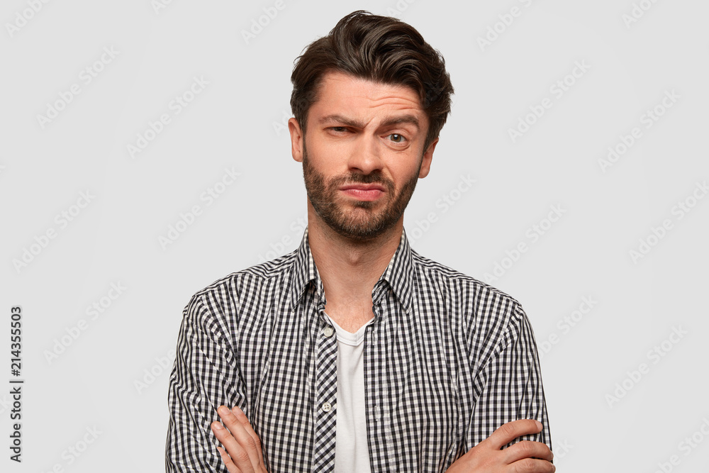 Stylish hipster male keeps arms folded, looks with hesitant expression, raises eyebrow with bewilderment, listens somebody`s suggestion, doubts whether to agree or not. Facial expressions concept Stylish hipster male keeps arms folded, looks with hesitant expression, raises eyebrow with bewilderment, listens somebody`s suggestion, doubts whether to agree or not. Facial expressions concept