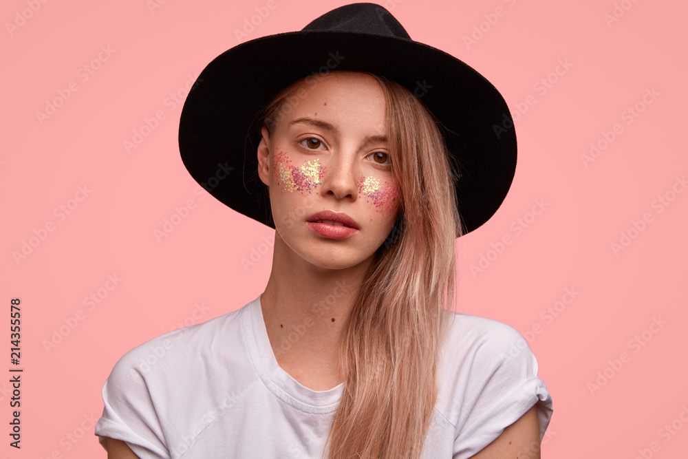 Attractive female hipster with bright sparkles on cheeks, wears fashionable black hat, casual white t shirt, stands against pink background, looks seriously at camera. People and beauty concept Attractive female hipster with bright sparkles on cheeks, wears fashionable black hat, casual white t shirt, stands against pink background, looks seriously at camera. People and beauty concept