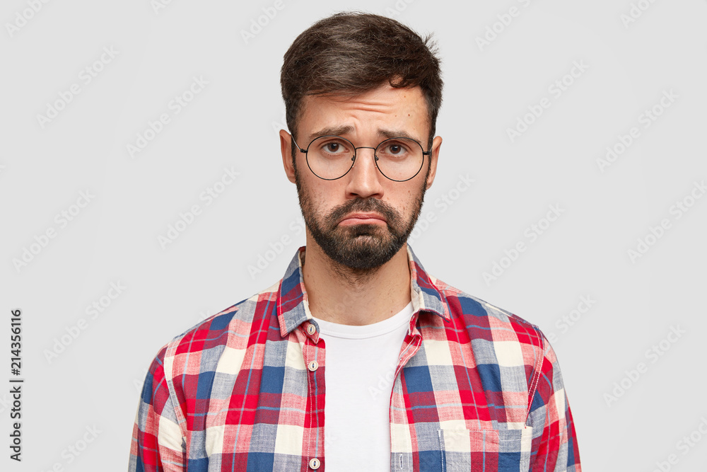 Upset young bearded male with gloomy expression, being offended by someone, raises eyebrows, needs support and help, wears glasses and casual checkered shirt, stands indoor, being in low spirit Upset young bearded male with gloomy expression, being offended by someone, raises eyebrows, needs support and help, wears glasses and casual checkered shirt, stands indoor, being in low spirit