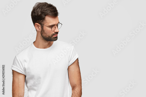 Photo of thoughtful Caucasian male with strong body shape, focused aside with pensive expression, contemplates about something in mind, wears casual t shirt, stands against white wall, free space © wayhome.studio 