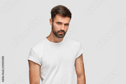 Portrait of serious bearded male hipster looks confidently at camera, has dark hair, wears casual t shirt, hears attentively something important, stands against white wall, has strong body shape
