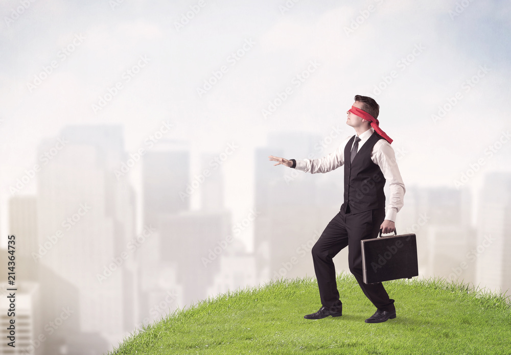Young blindfolded businessman steps on a a patch of grass with a city in the background Young blindfolded businessman steps on a a patch of grass with a city in the background