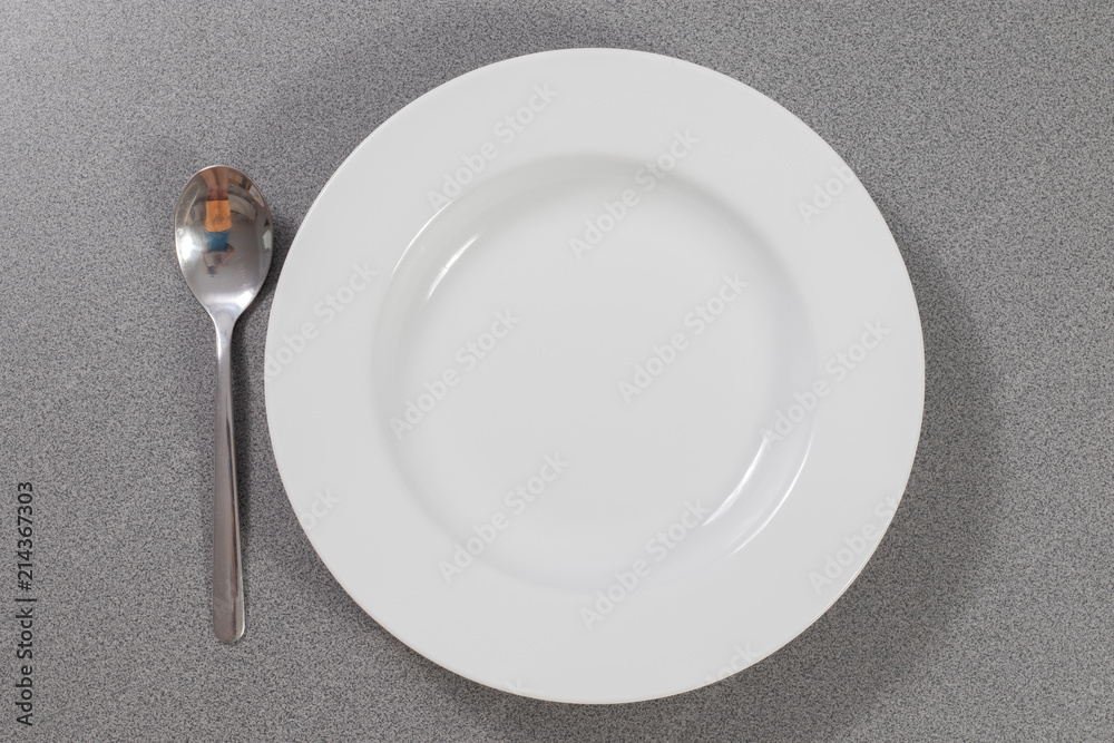Suppenlöffel aus Metall neben einem weißen Suppenteller auf einem grauen Tisch Suppenlöffel aus Metall neben einem weißen Suppenteller auf einem grauen Tisch