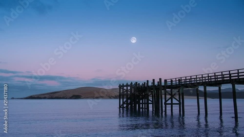 View across calm lapping water in Hokianga Harbour at dawn with full moon rising over wooden pier. New Zealand. photo