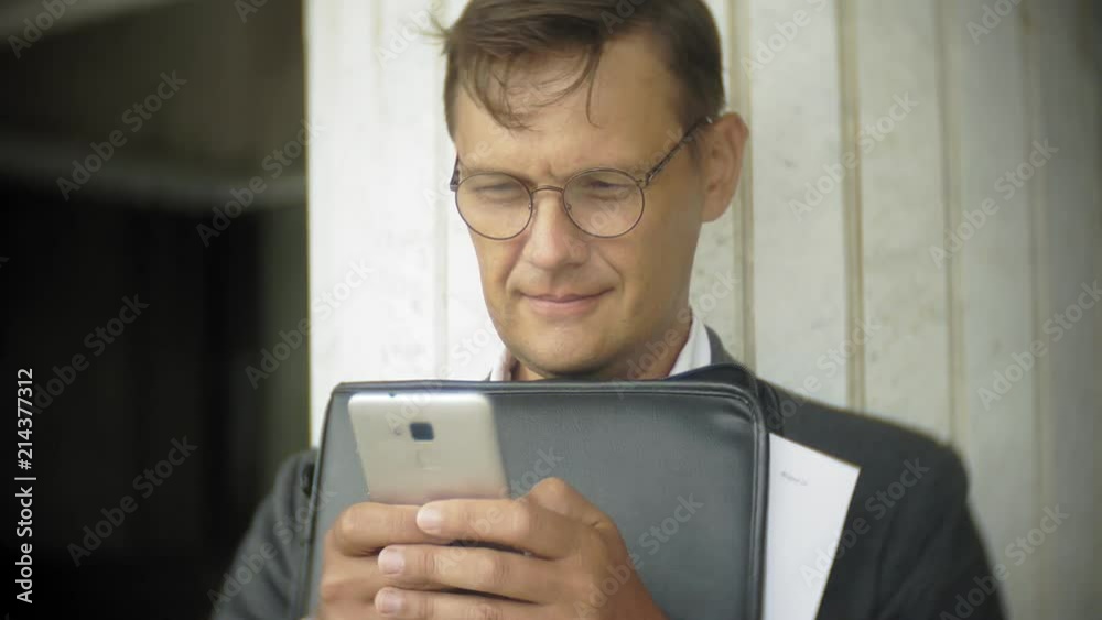 Businessman is sitting on the stairs in the city. He wears a suit and briefcase. He looks through documents and talks on the smartphone Businessman is sitting on the stairs in the city. He wears a suit and briefcase. He looks through documents and talks on the smartphone