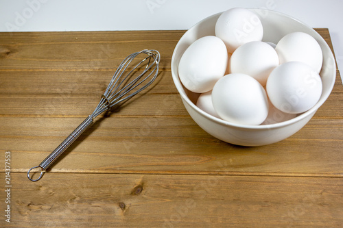 A group of eggs inside a deep white bowl next to a whisk waiting for the chef to use them in a meal