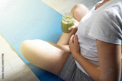 Pregnant woman drinking healthy green smoothie while sitting on yoga blue mat after exercise. Healthy pregnancy