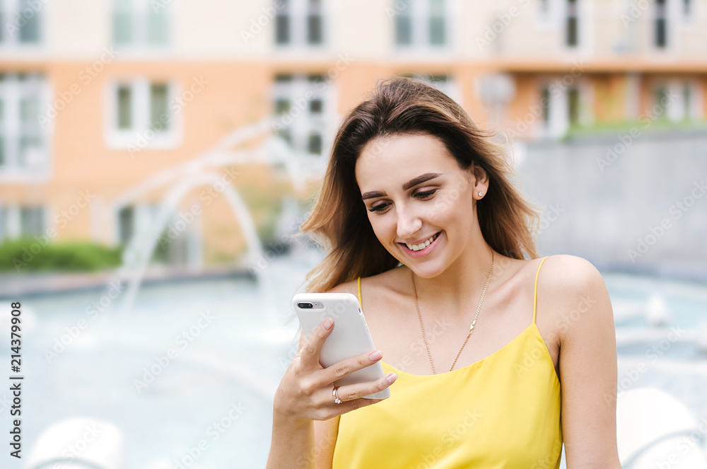Pretty young girl using mobile phone. Woman reading message, sms on smartphone, smiling, happy. Social media, communication people, gadget concept. Outdoor close up photo.  Multistory house on back
