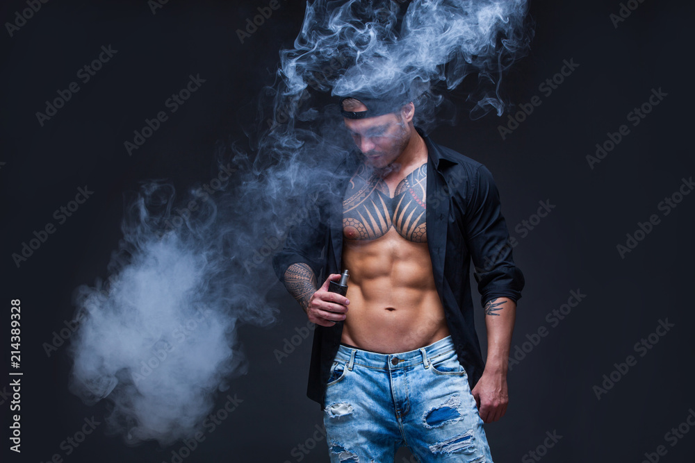 Vaper. The man dressed blue jeans, black shirt and black baseball cap with tattoos smoke an electronic cigarette on the dark background Vaper. The man dressed blue jeans, black shirt and black baseball cap with tattoos smoke an electronic cigarette on the dark background
