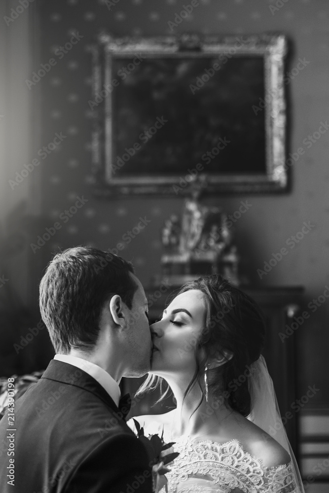 gorgeous bride with bouquet and stylish groom gently kissing in luxury room in hotel. rich wedding couple embracing. romantic sensual moment of newlyweds in amazing classic indoors gorgeous bride with bouquet and stylish groom gently kissing in luxury room in hotel. rich wedding couple embracing. romantic sensual moment of newlyweds in amazing classic indoors