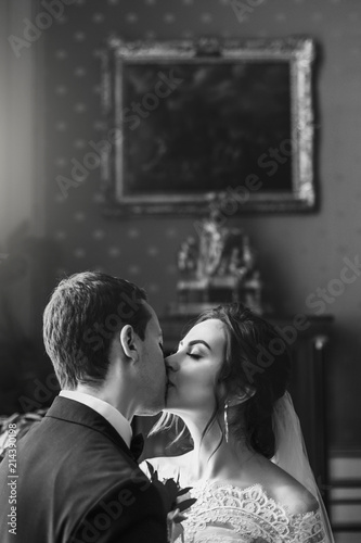 gorgeous bride with bouquet and stylish groom gently kissing in luxury room in hotel. rich wedding couple embracing. romantic sensual moment of newlyweds in amazing classic indoors