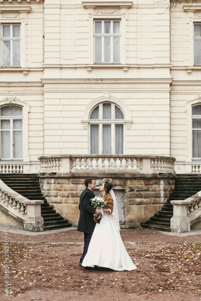 Obraz premium gorgeous bride with bouquet and stylish groom walking at old castle in european city street in autumn time. happy wedding couple embracing. romantic sensual moment of newlyweds