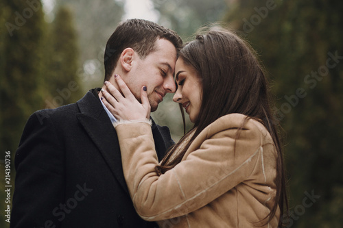 gorgeous wedding couple kissing in winter snowy park. stylish bride in coat and groom embracing under green trees in winter forest. romantic sensual moment of newlyweds