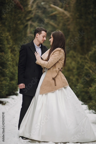 gorgeous wedding couple posing in winter snowy park. stylish bride in coat and  groom embracing under green trees in winter forest. romantic sensual moment of newlyweds