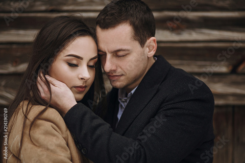gorgeous bride in coat and stylish groom posing at wooden house in winter forest. happy wedding couple gently hugging in snowy park.  barn wedding. romantic sensual moment