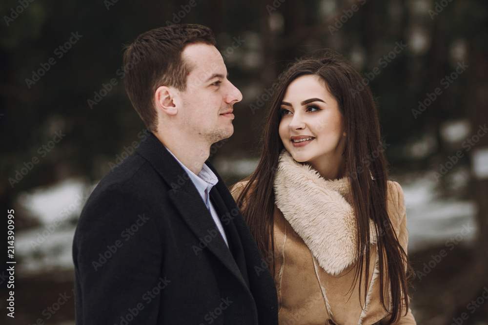 Fototapeta premium gorgeous wedding couple walking in winter snowy park. stylish bride in coat and groom embracing under green trees in winter forest. romantic sensual moment of newlyweds