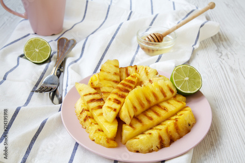 Grilled pineapple wedges on pink plate with lime and honey over white wooden surface, side view. Summer food. Idea for snack. Closeup.