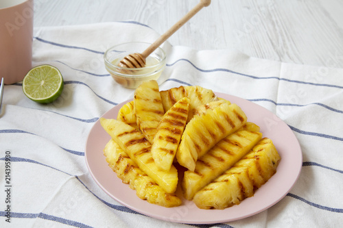 Grilled pineapple wedges on pink plate with lime and honey over white wooden background, side view. Summer food. Close-up.
