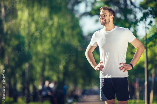 Full length of smiling attractive athlete standing and holding hands on waist. He is enjoying perfect weather for outdoor training. Copy space in left side