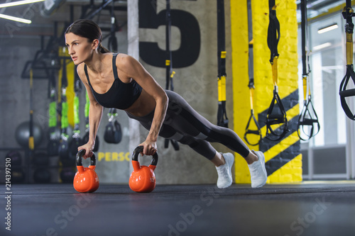 Athletic woman is straining in plank position. She is having hands on heavy weights and looking ahead. Female is doing crossfit training with equipment in fitness center