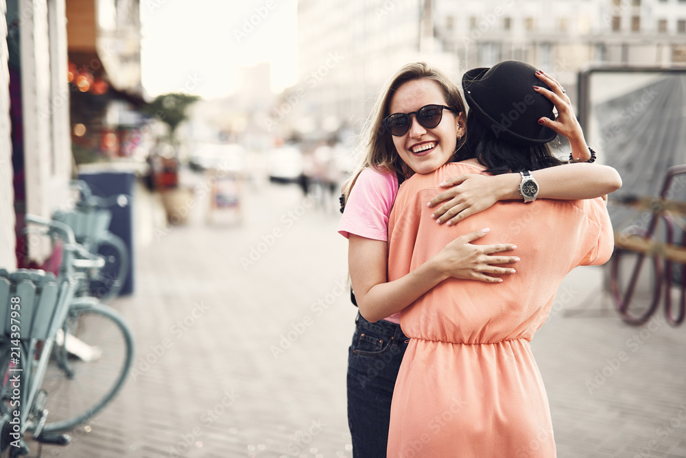 Obraz premium Positive girl hugging female friend. They standing on street during sunny day