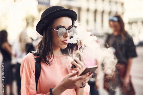 Portrait of orderly young lady smoking electronic cigarette while using contemporary phone outside photo