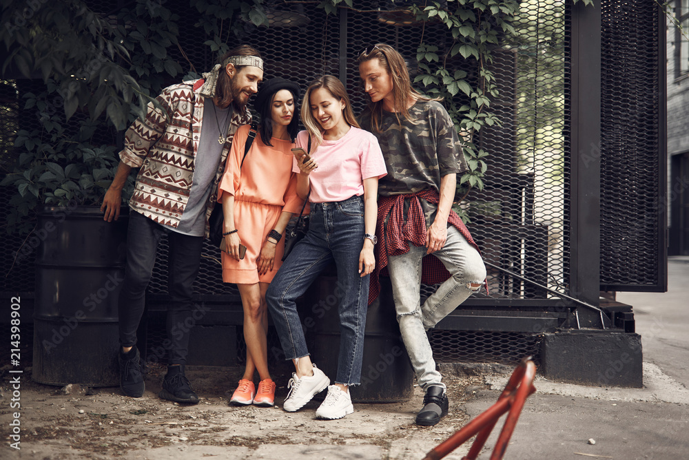 Fototapeta premium Full length outgoing female holding modern cellphone in arm while situating near cheerful friends. Satisfied ladies and glad males looking at mobile outdoor