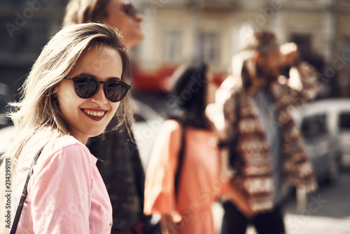 Portrait of cheerful girl with beautiful smile wearing modern sunglasses. She looking at camera while walking with friends along street