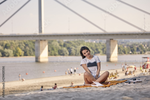 one young beautiful woman smiling, sunny day, reading a book, looking to camera, sitting on blanket, sandy beach. Unrecognizable people in background. Coast, bridge, river in background.