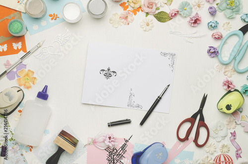 A card in the center on a white background surrounded by colored paper, paper flowers and scrapbooking materials. Top view