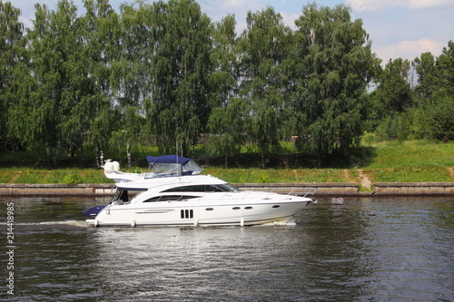 Travel, luxury water recreation on boat - a white 58-ft motor yacht slowly sails along the concrete shores of the navigable canal in summer amid the green trees on the shore, side view