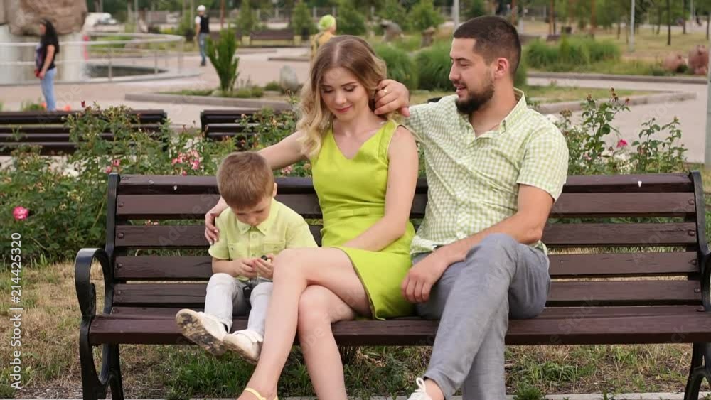 Happy young family with his son resting in the Park in the summer on a bench. A beautiful woman with her husband and a child sitting on a bench in the background of the urban landscape. Happy young family with his son resting in the Park in the summer on a bench. A beautiful woman with her husband and a child sitting on a bench in the background of the urban landscape.