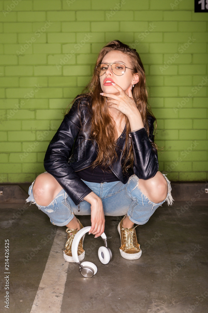 Fashion portrait of beautiful woman DJ in cool glasses, rock black leather  jacket slav squat sit and with white music headphones sound. Hipster urban  style girl. Lifestyle outdoor city portrait. Photos