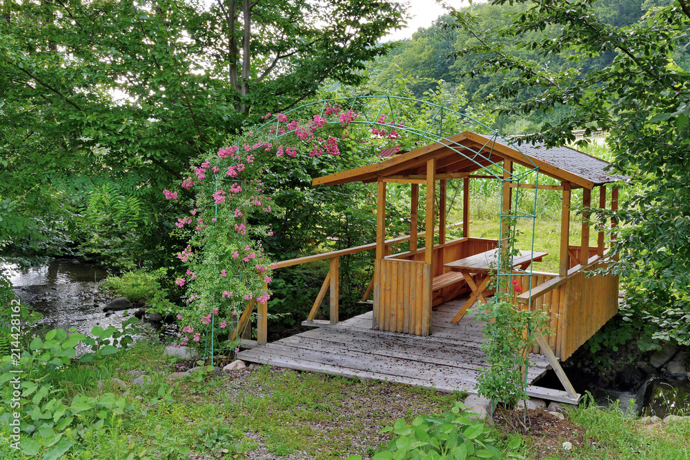 Wooden gazebo with a roof and a table standing on the boards lying over the stream below. And growing flowers on an arch of wire made nearby. Wooden gazebo with a roof and a table standing on the boards lying over the stream below. And growing flowers on an arch of wire made nearby.