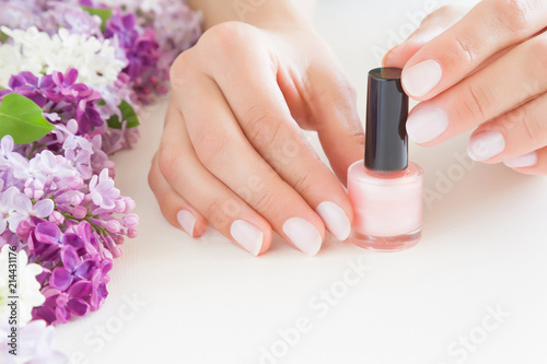 Young  perfect  groomed woman s hands with pink nail varnish bottle. Nails care. Manicure  pedicure beauty salon. Beautiful branches of lilac blossoms on white table. Colorful  fresh flowers.