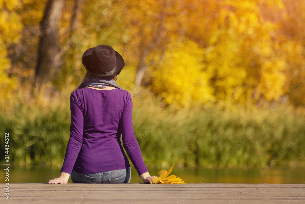 Girl in a hat sits on the dock and admires the colors of autumn. Back view