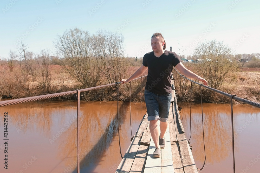 Naklejka premium Young man is walking on a suspended wooden bridge over the river.