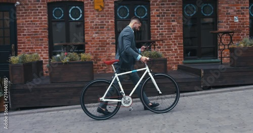 TRACKING Handsome young adult man wearing suit choosing music on his phonee before riding his classic bicycle to work in the morning. 4K UHD 60 FPS SLOW MOTION photo