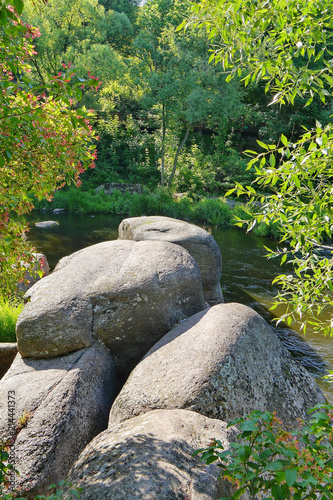 Large stones with water-polished surface lying on the shore of a small river amidst picturesque nature in green thickets of bushes and trees.