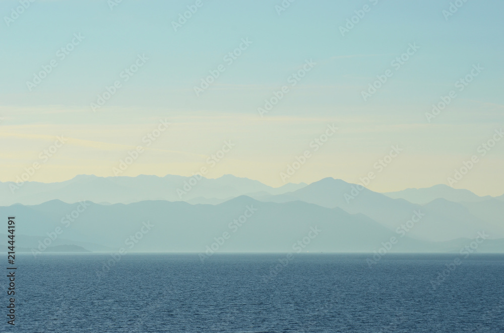 A series of blue hills run down to a blue ocean. Above is a pale blue sky with white clouds. A series of blue hills run down to a blue ocean. Above is a pale blue sky with white clouds.