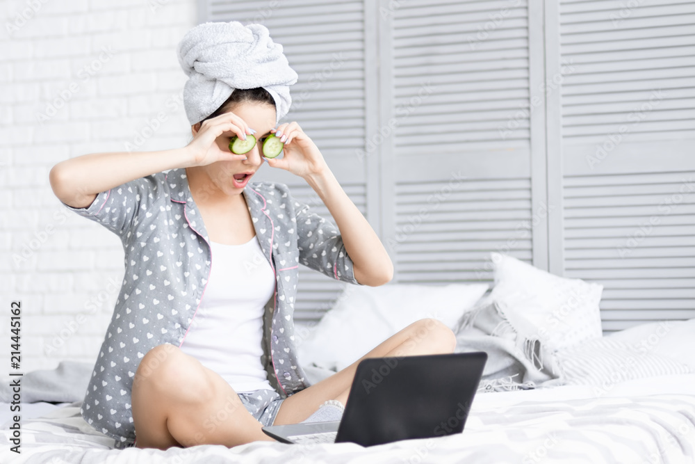 Young girl woman female adult after shower wearing pajamas and towel over her head sitting in her bed with cucumber slices holding in front of her eyes and surprised looking at laptop. Daily routine Young girl woman female adult after shower wearing pajamas and towel over her head sitting in her bed with cucumber slices holding in front of her eyes and surprised looking at laptop. Daily routine